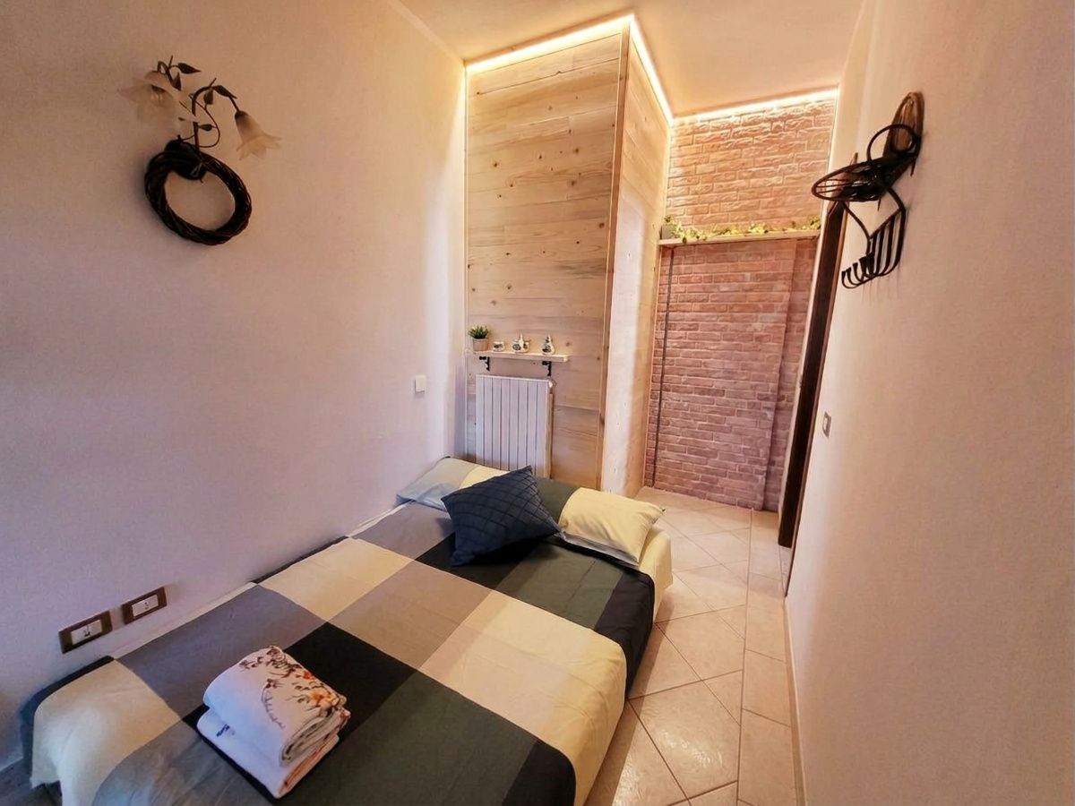 Une petite chambre avec une couvre-lit à carreaux, une table de chevet, un luminaire fixé au mur et une vue sur un mur de briques.