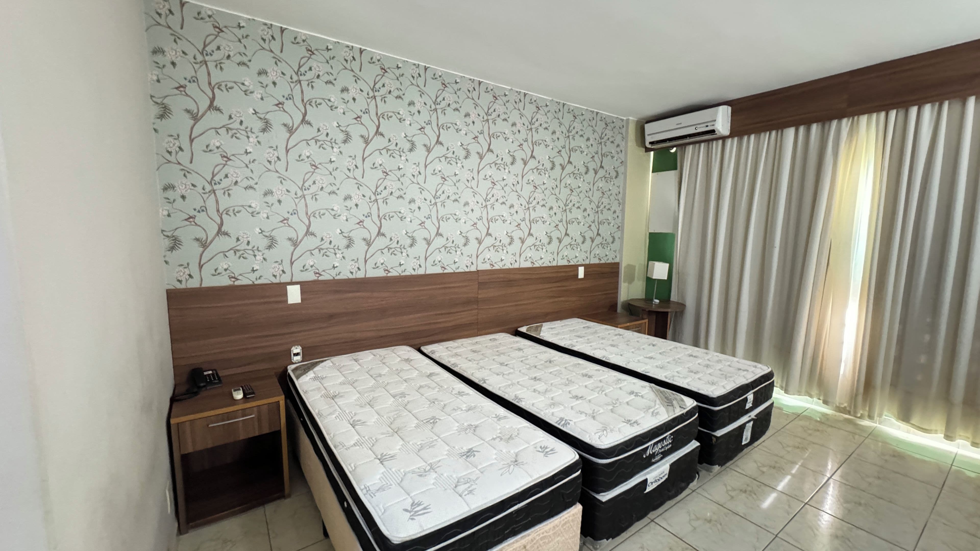 Quarto de hotel com duas camas, cabeceira de madeira, papel de parede floral, ar condicionado, mesa de cabeceira com abajur e janela com cortinas.