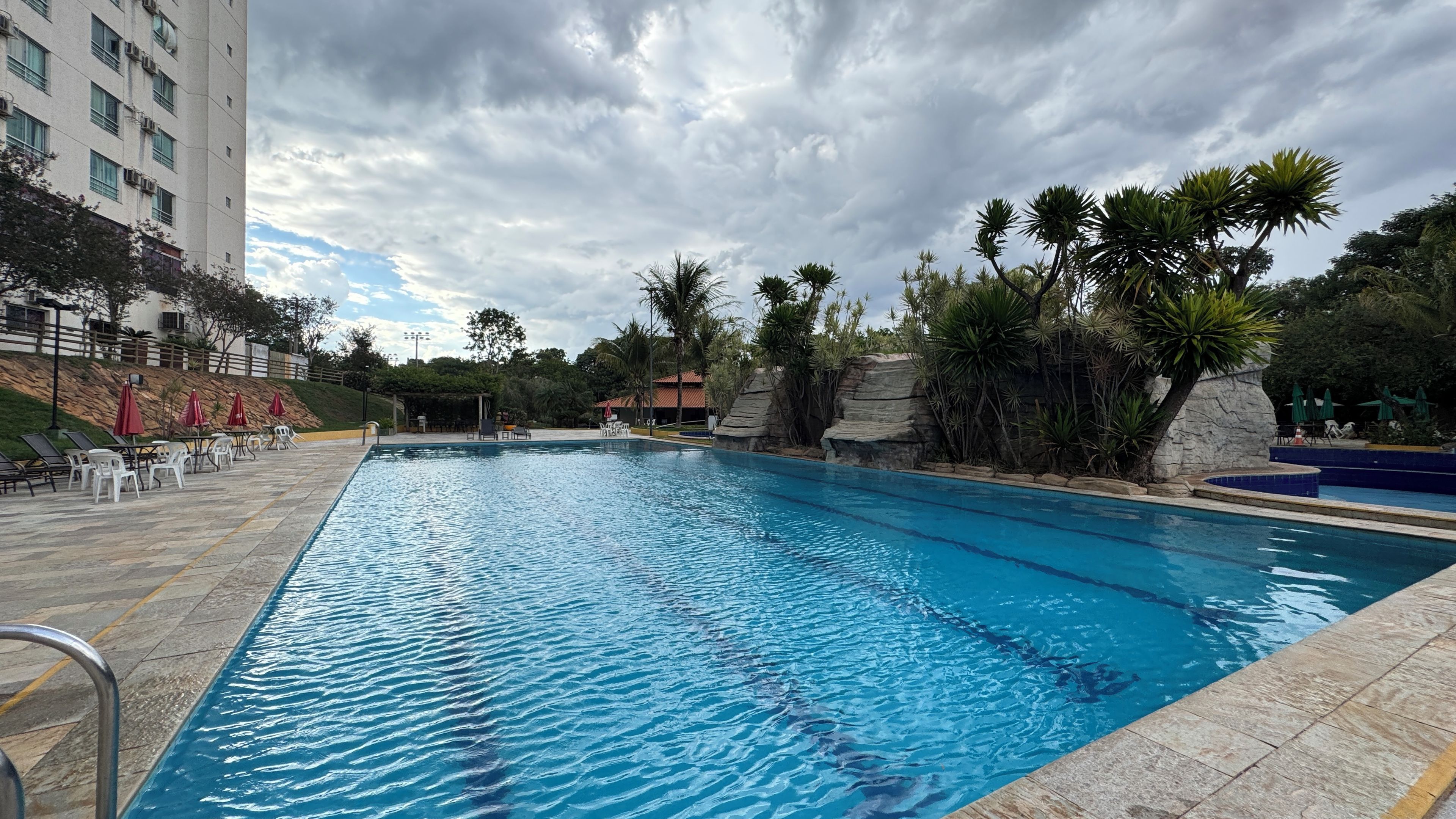 A swimming pool with clear water, surrounded by lounge chairs and umbrellas, with a building and trees in the background.