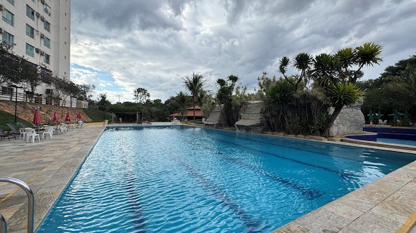 A swimming pool with clear water, surrounded by lounge chairs and umbrellas, with a building and trees in the background.