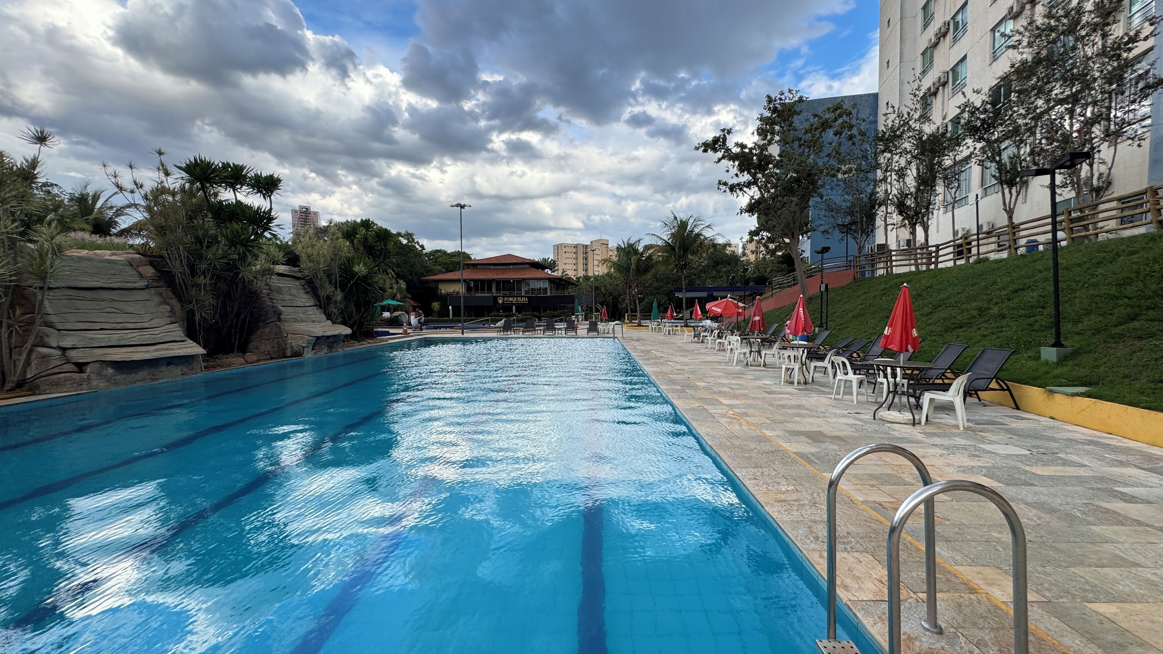 A swimming pool with lounge chairs and umbrellas, surrounded by greenery and buildings.
