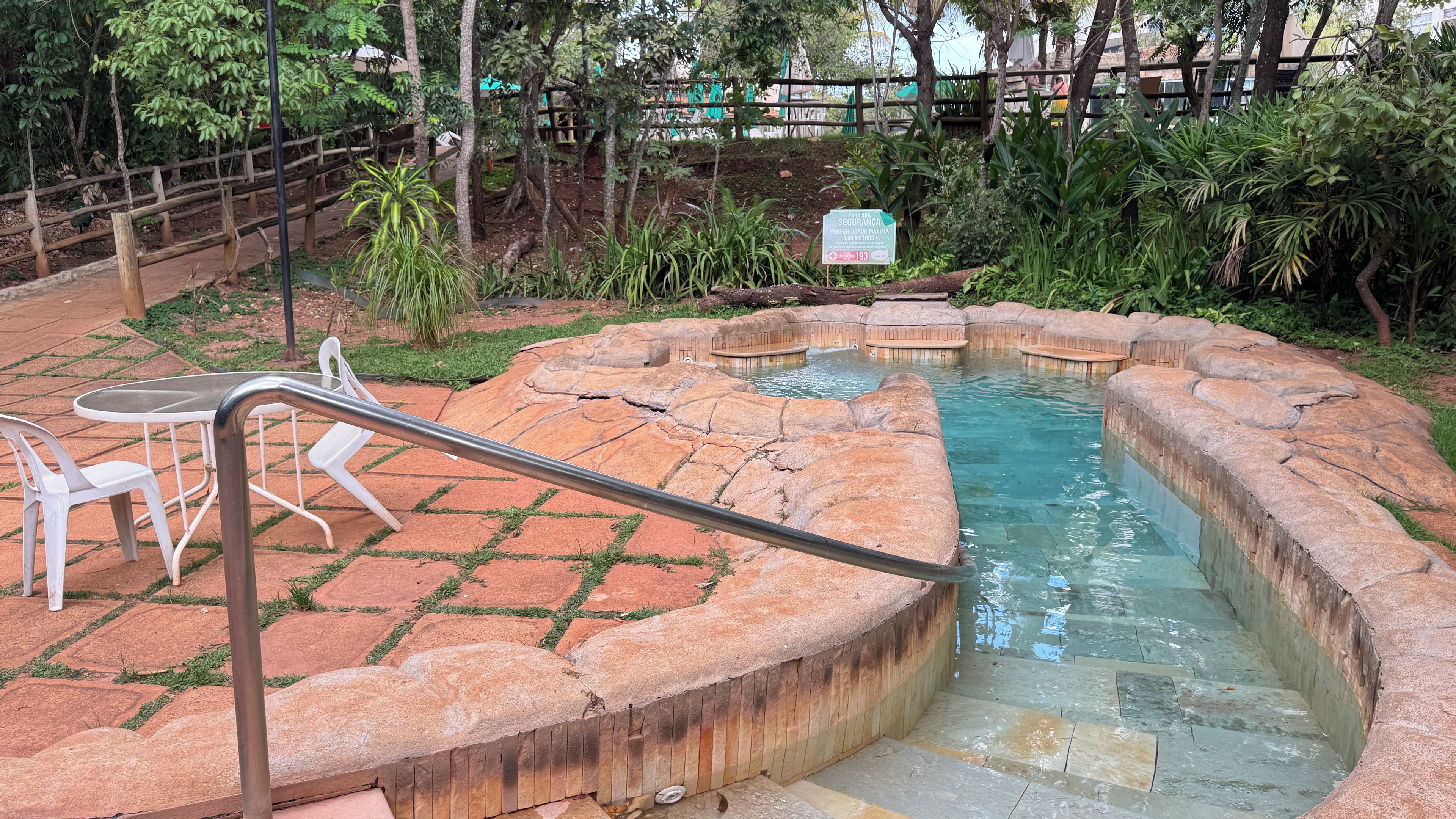 A hot spring area with a wooden bench and a table, surrounded by a wooden fence and lush greenery.