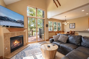 A spacious living room with a fireplace, a leather sofa, a television, and a dining area with natural light. - Highland Greens Townhome: Long-Stay Lodging, Private Hot Tub (Breckenridge)