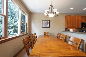 Une salle à manger avec une longue table en bois et des chaises, un lustre et une fenêtre donnant sur les arbres.