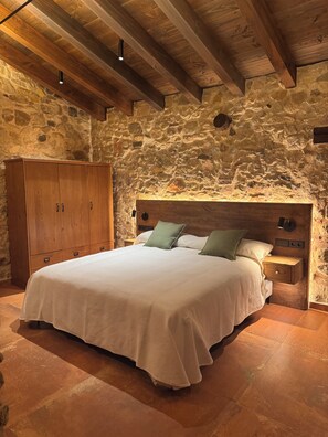 A bedroom with a stone wall, wooden ceiling beams, a large bed, a wooden wardrobe, and a nightstand with a lamp. - Can Roig Farmhouse (Riells i Viabrea)