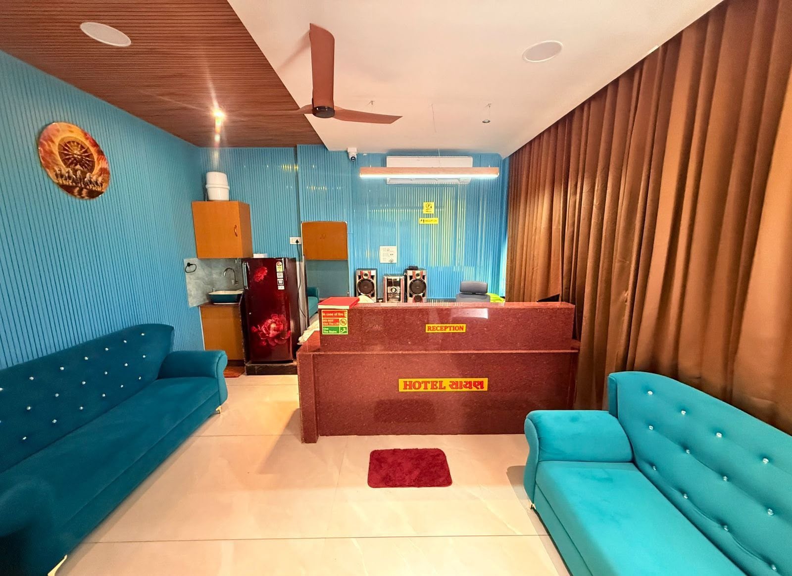 Zona reception di un hotel con scrivania, due divani blu, un frigorifero rosso e una ventola a soffitto.