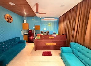 A hotel reception area with a reception desk, two blue sofas, a red refrigerator, and a ceiling fan.