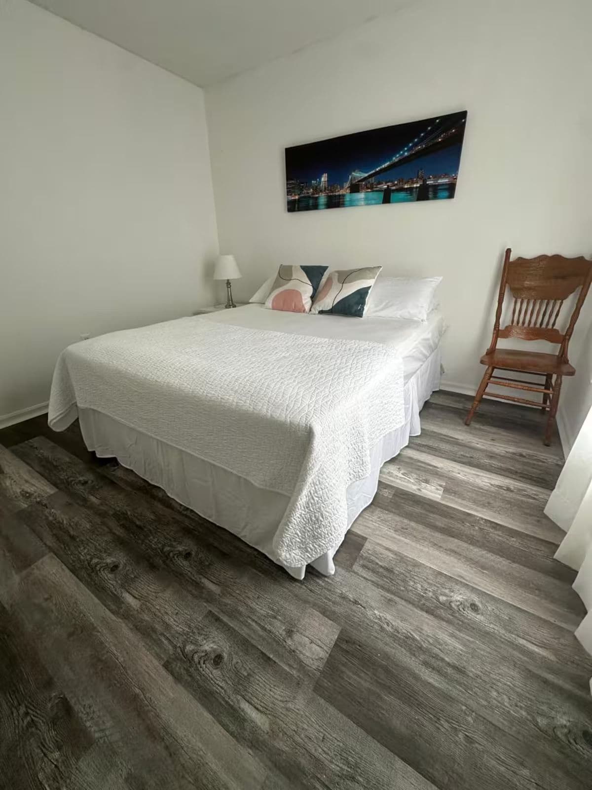A bedroom with a large bed, a chair, a nightstand, a lamp, and a cityscape painting on the wall.