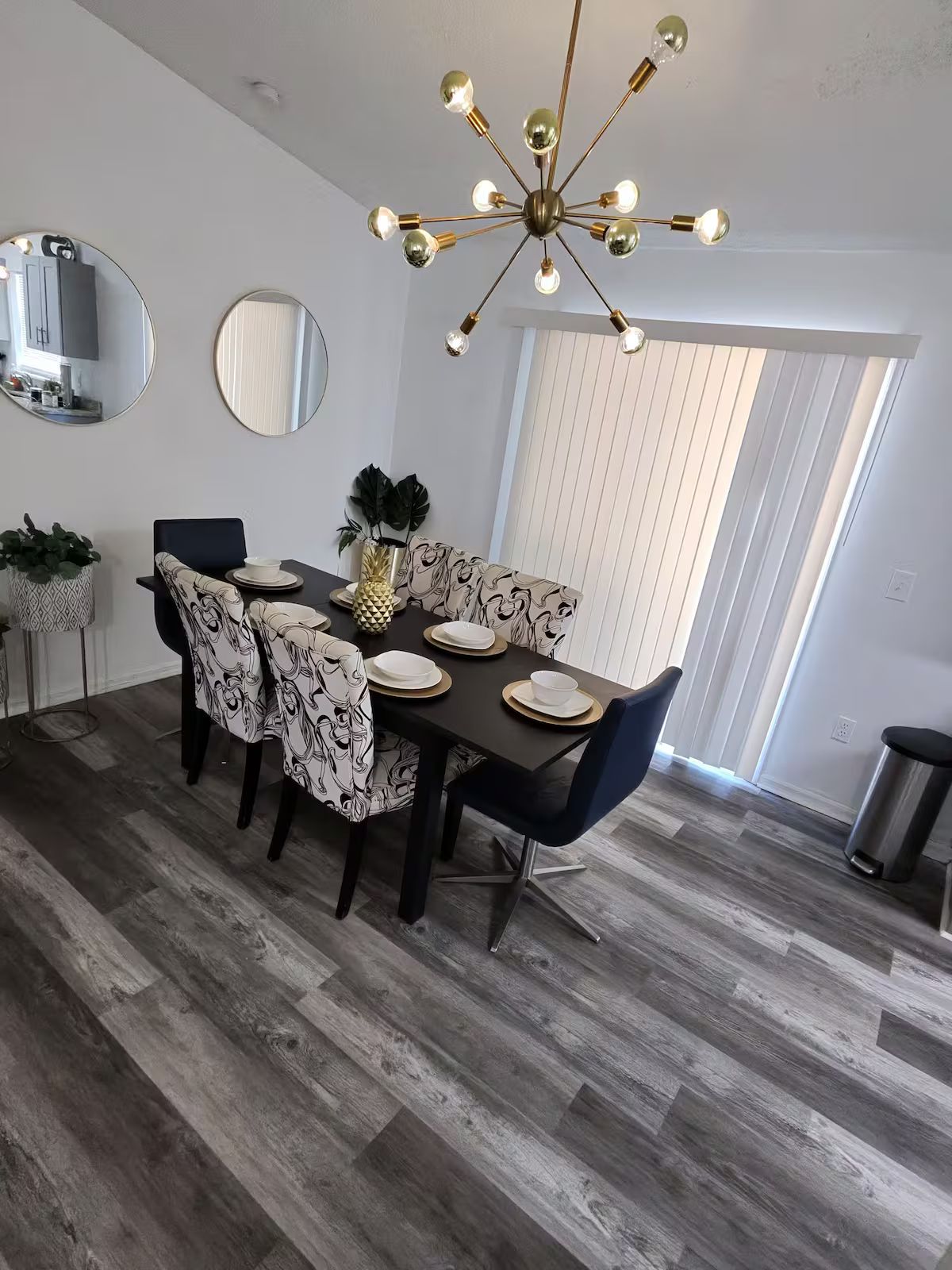 A dining area with a modern chandelier, round mirrors, and a large window with blinds.
