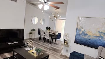 Un salon moderne doté d’un téléviseur à écran plat, d’un ventilateur de plafond, d’un coin repas avec une table et des chaises, et d’un grand tableau abstrait accroché au mur.