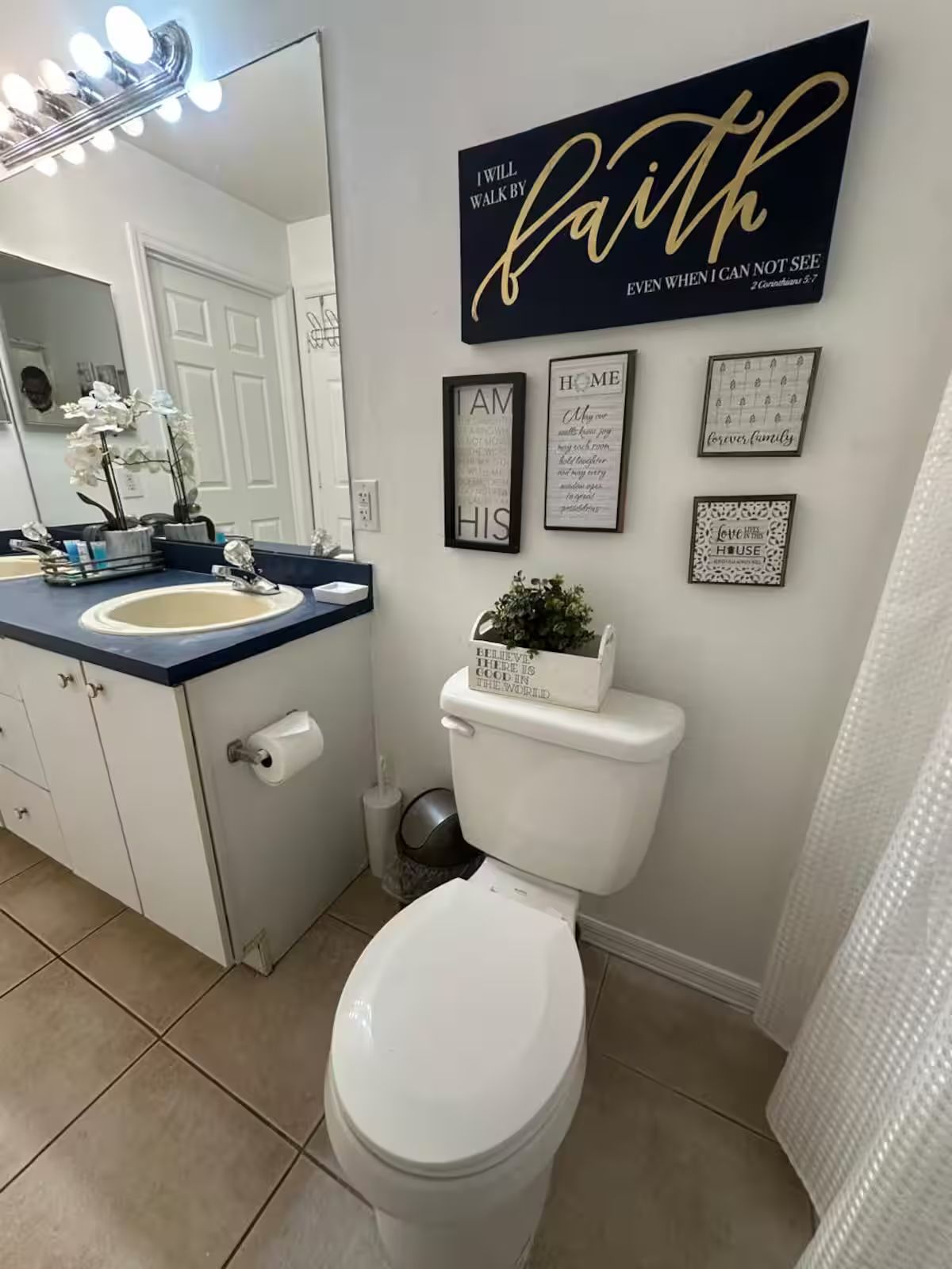 A bathroom with a white toilet, a sink with a blue countertop, and a mirror with lights. There are framed inspirational quotes on the wall.