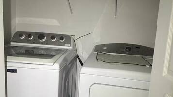 A laundry room with two washing machines and a shelf above containing a water bottle, a red container, and a box labeled 'DRYER SHEETS'.