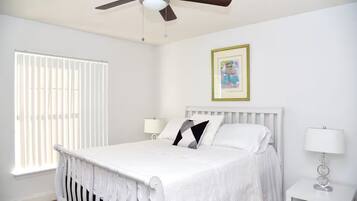 A bedroom with a bed, a ceiling fan, a window with blinds, a bedside table with a lamp, and a framed picture on the wall.