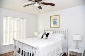 A bedroom with a bed, a ceiling fan, a window with blinds, a bedside table with a lamp, and a framed picture on the wall.