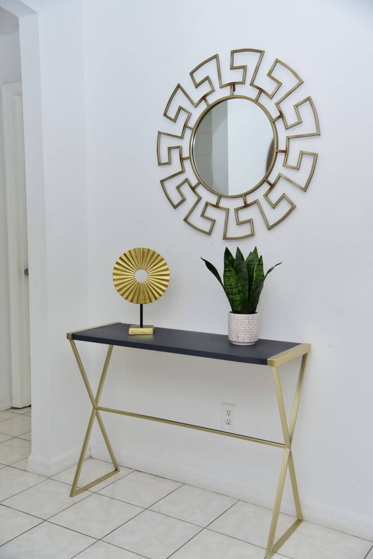 A white wall with a gold-framed mirror, a small table with a gold frame, a gold-framed lamp, and a potted plant.
