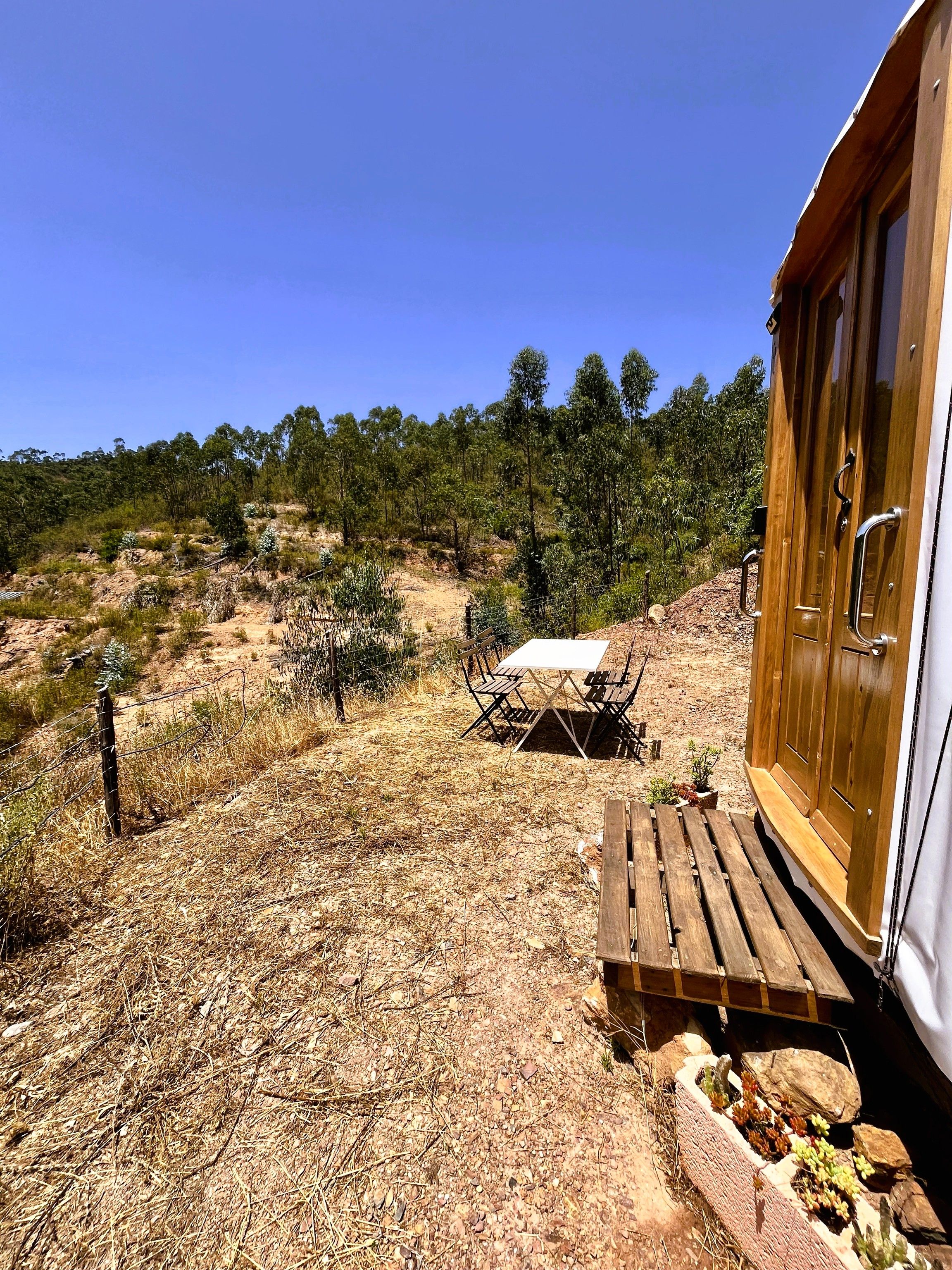 A wooden cabin with a table and chairs outside, surrounded by dry grass and sparse vegetation.