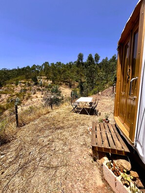 A wooden cabin with a table and chairs outside, surrounded by dry grass and sparse vegetation.