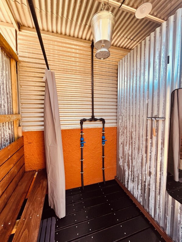 A compact, enclosed space with corrugated metal walls, a wooden bench, a hanging light fixture, and a black floor with small reflective dots.