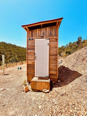 A wooden outhouse with a door, number 2, situated on a gravel path with a forested hill in the background.