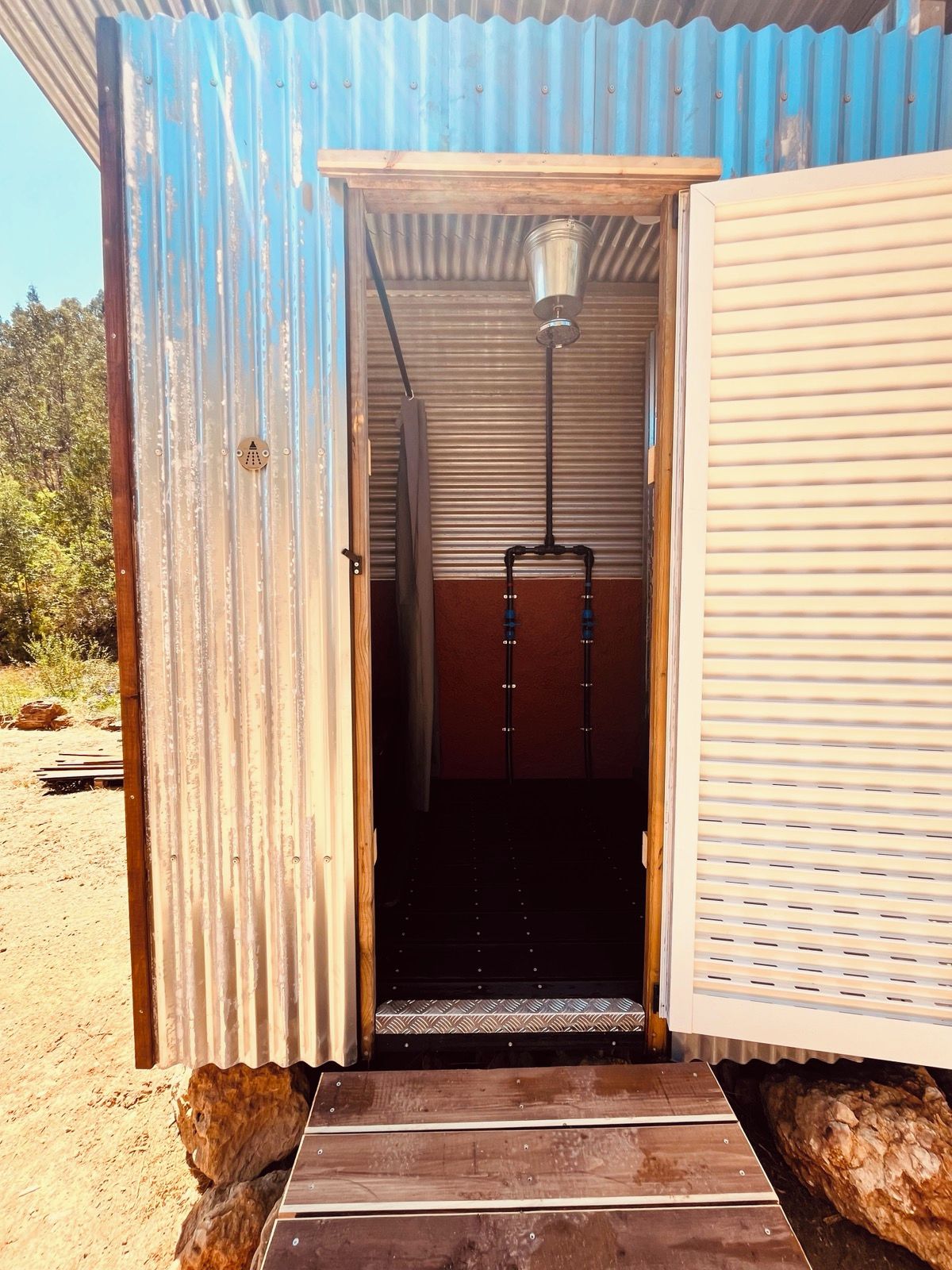 A small, open-sided structure with a metal roof and a wooden door. Inside, there is a black bench and a metal bucket hanging from the ceiling.