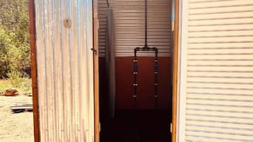 A small, open-sided structure with a metal roof and a wooden door. Inside, there is a black bench and a metal bucket hanging from the ceiling.