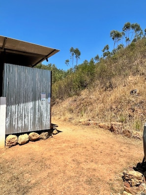 Un edificio de metal corrugado con techo plano, ubicado en un sendero de tierra con rocas al frente.