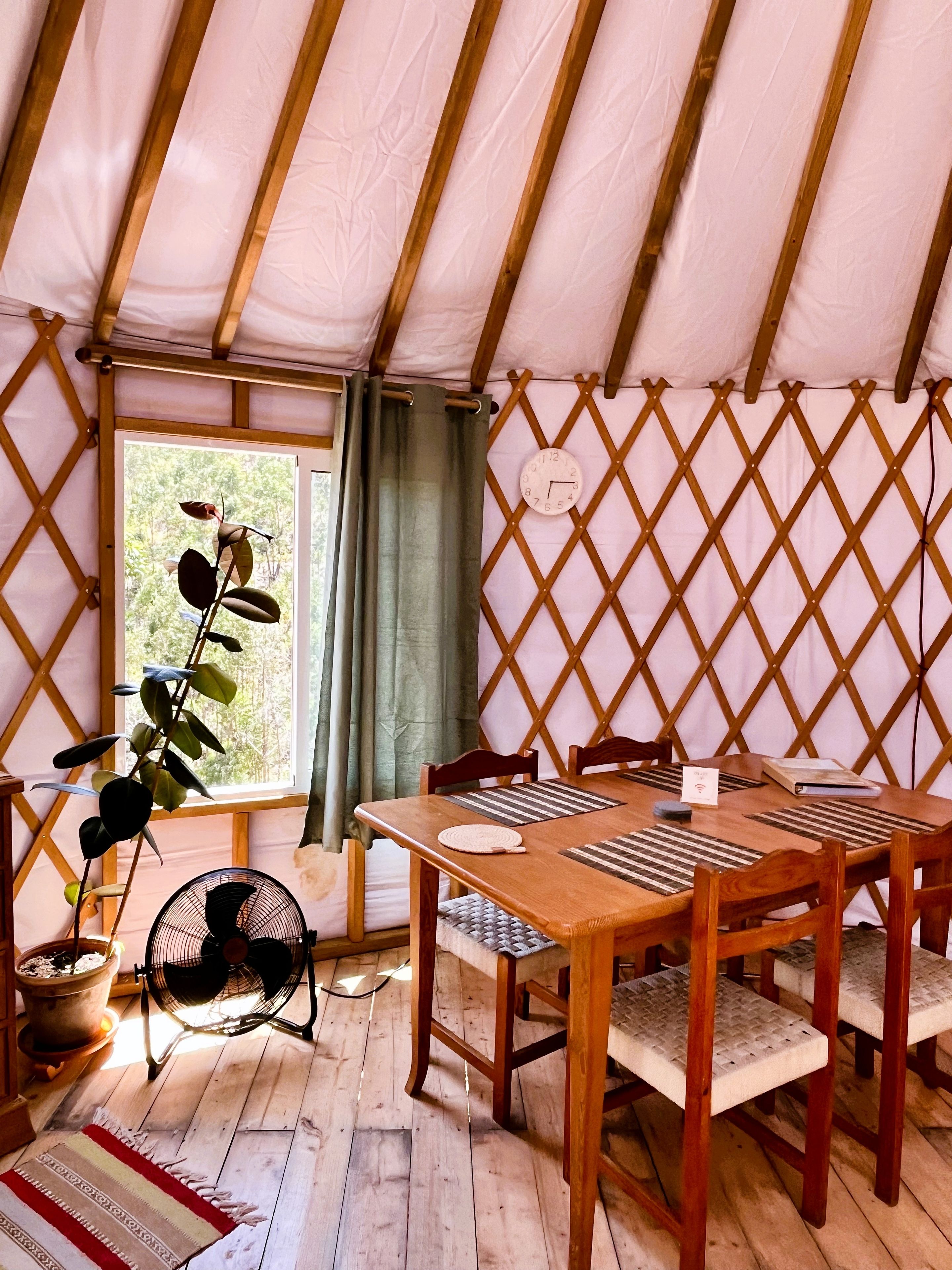 A tent with a wooden table and chairs, a fan, and a plant.