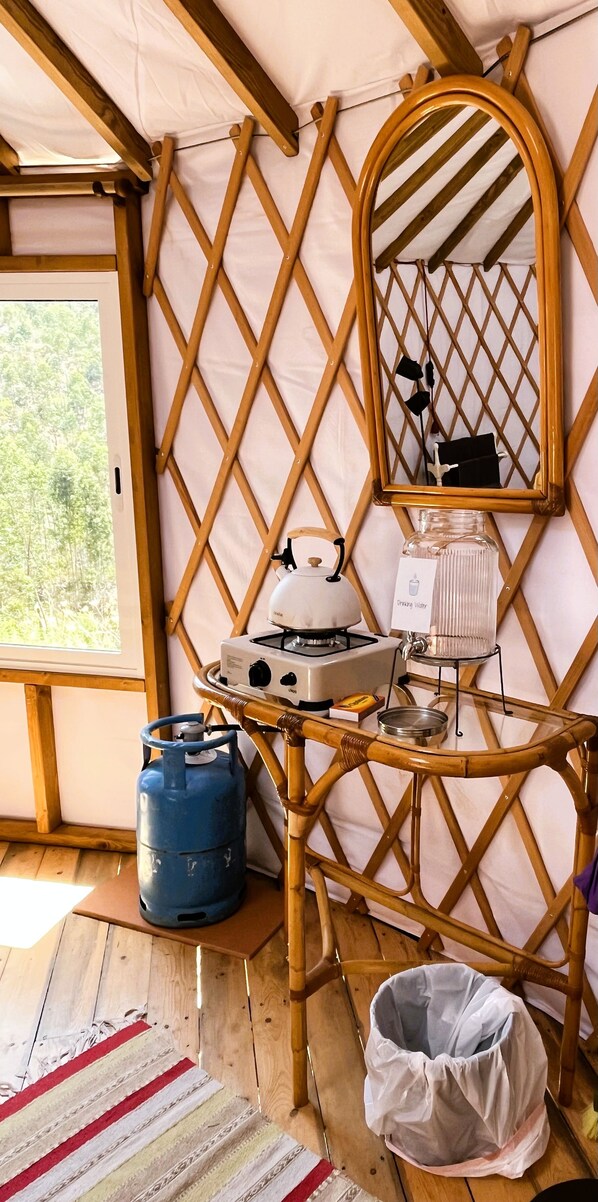 Una habitación con una mesa de bambú, estufa a gas, hervidor, bombona de gas azul y cubo de basura blanco.