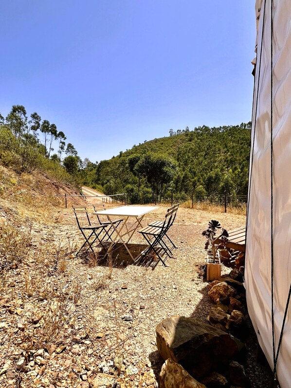 A campsite with a table and chairs set up on a gravel surface, surrounded by dry vegetation and a hilly backdrop.
