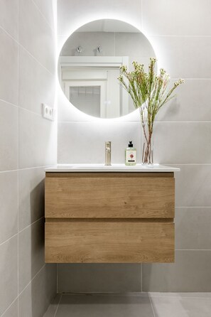 A bathroom with a wooden vanity, a round mirror, and a vase with flowers. - Newly renovated in the heart of Madrid (Madrid)