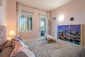 A bedroom with a bed, a nightstand, a lamp, a window with curtains, a door, and a cityscape painting on the wall. - Cubo's Opalo de Riviera Apartment / communal pool / communal gym / air conditioning / terrace (Mijas Costa)