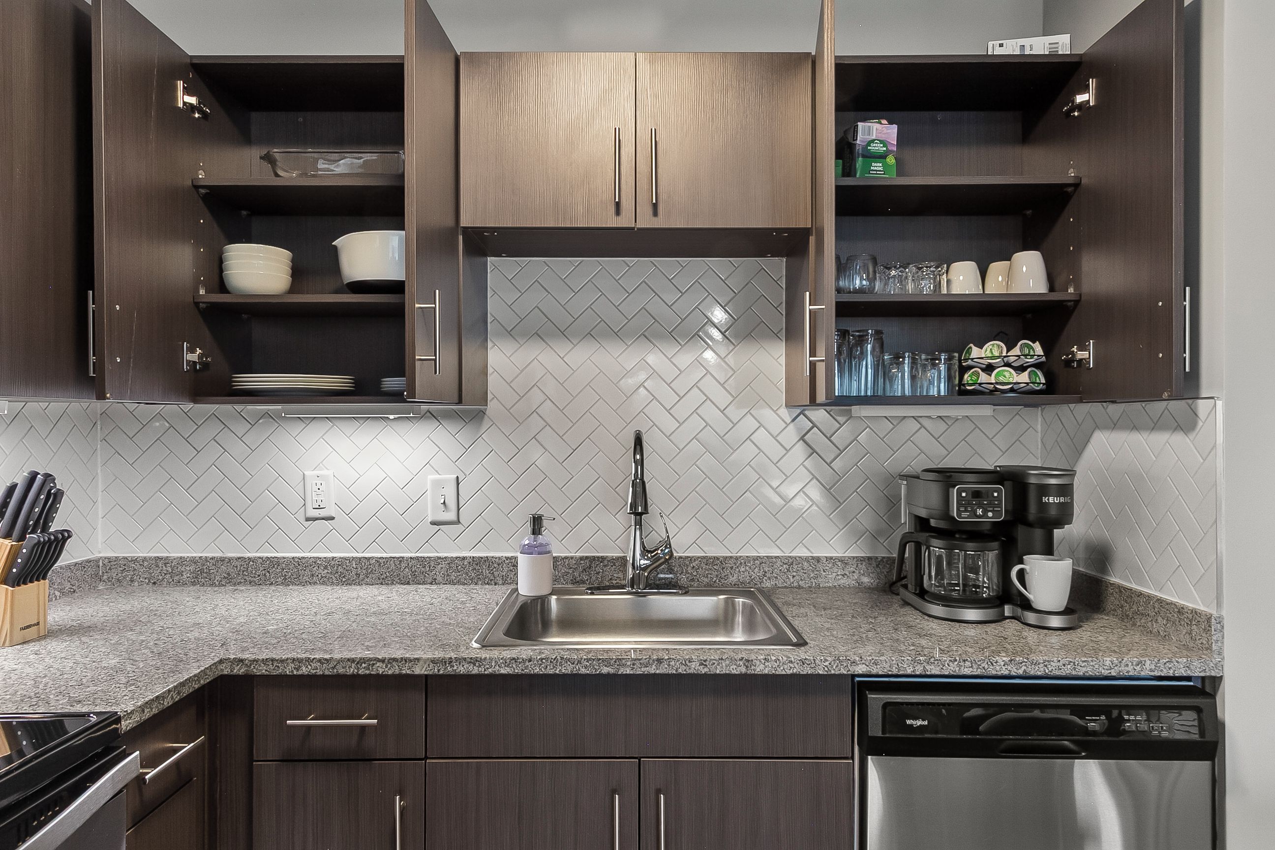Een moderne keuken met donkere houtkasten, een backsplash van mozaïektegels en een roestvrijstalen spoelbak, en een koffiezetapparaat.