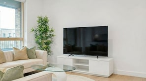 A modern living room with a flat-screen TV on a white stand, a glass coffee table, a beige sofa with cushions, and a tall green plant. - 2-Bed 2-Bath London Apartment, Fast Transport Links - Business & Long Stays (LONDON)