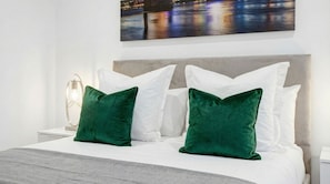 Ein ordentlich bezogenes Bett mit weißen und grünen Kissen, einem grauen Kopfteil und einer modernen Nachttischlampe.