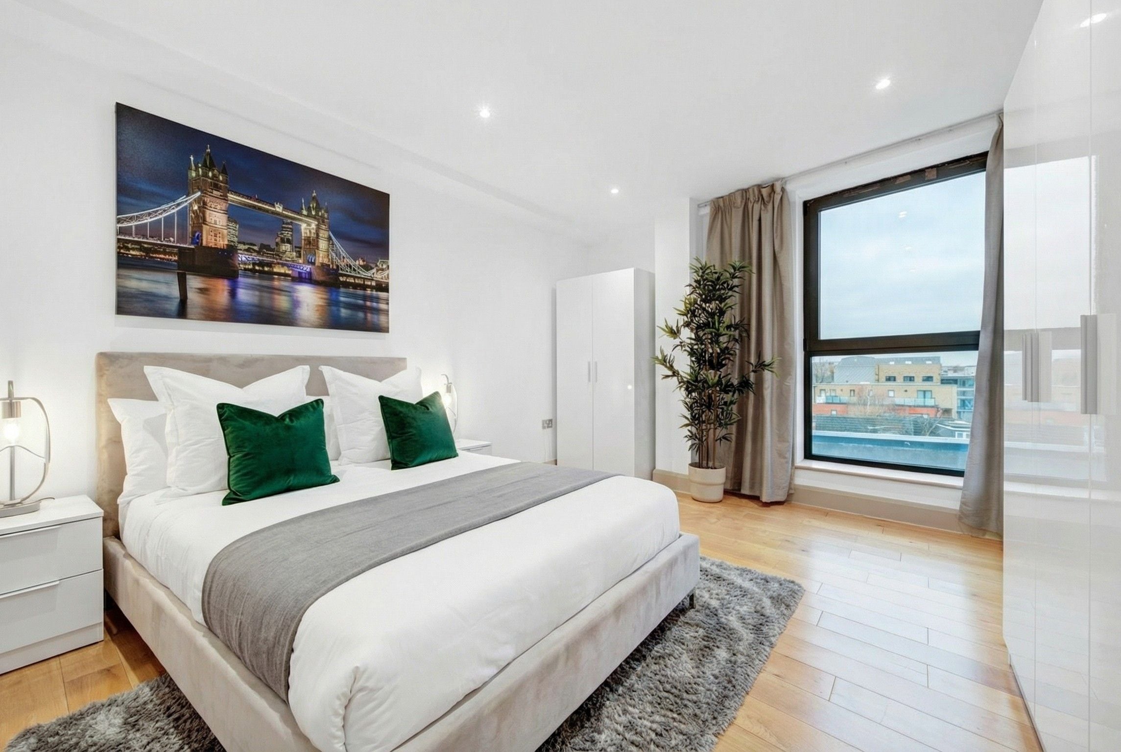 Un dormitorio moderno con una cama grande, una mesita de noche, una lámpara de noche, un ventanal con vistas y un cuadro de un puente.