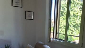 Ein Wohnzimmer mit einem beigefarbenen Sofa, zwei gerahmten Bildern an der Wand und einem Fenster mit Blick ins Grüne.