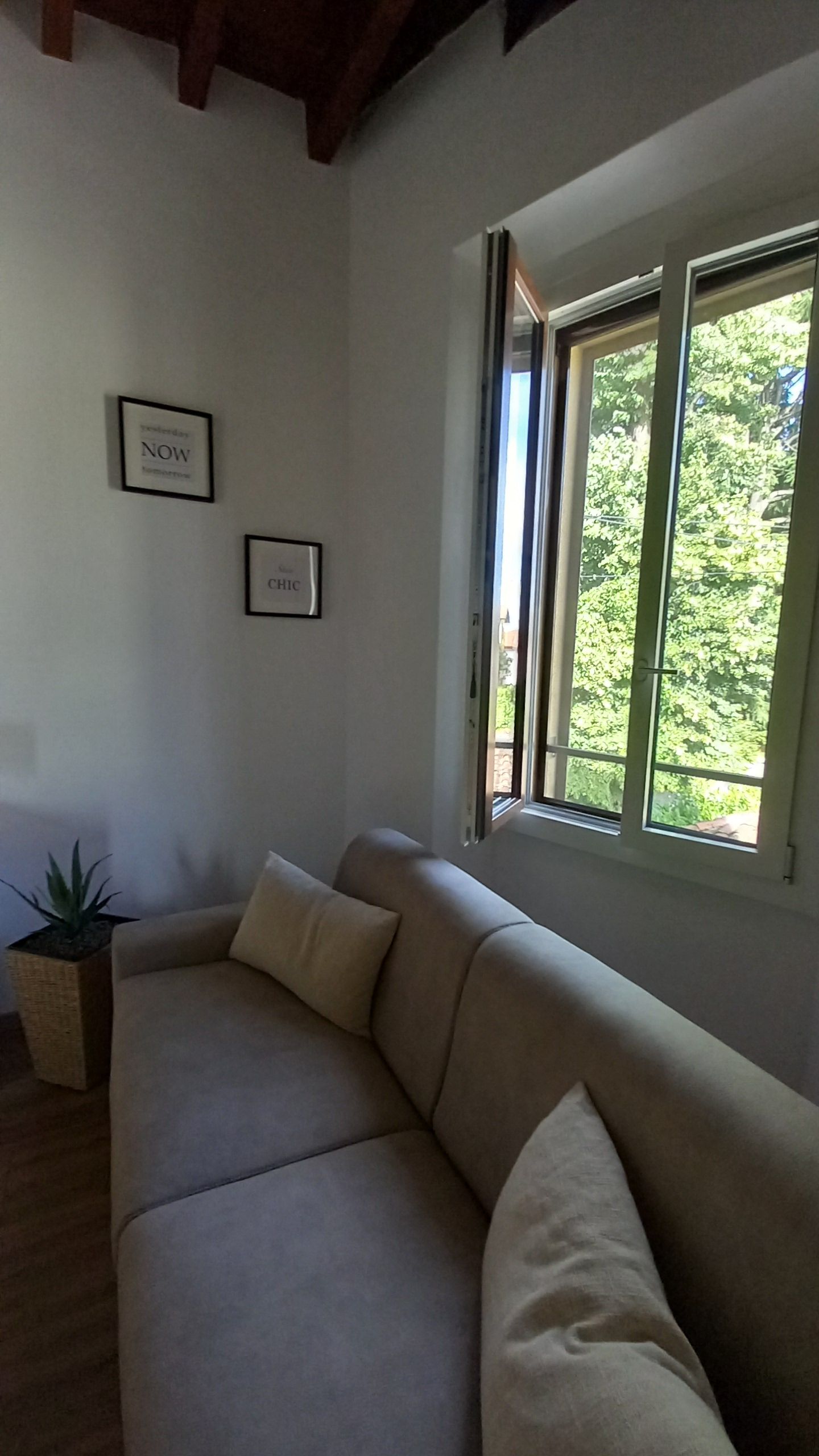 Een woonkamer met een beige bank, twee ingelijste foto's aan de muur en een raam met uitzicht op groen.