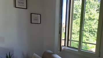 Een woonkamer met een beige bank, twee ingelijste foto's aan de muur en een raam met uitzicht op groen.