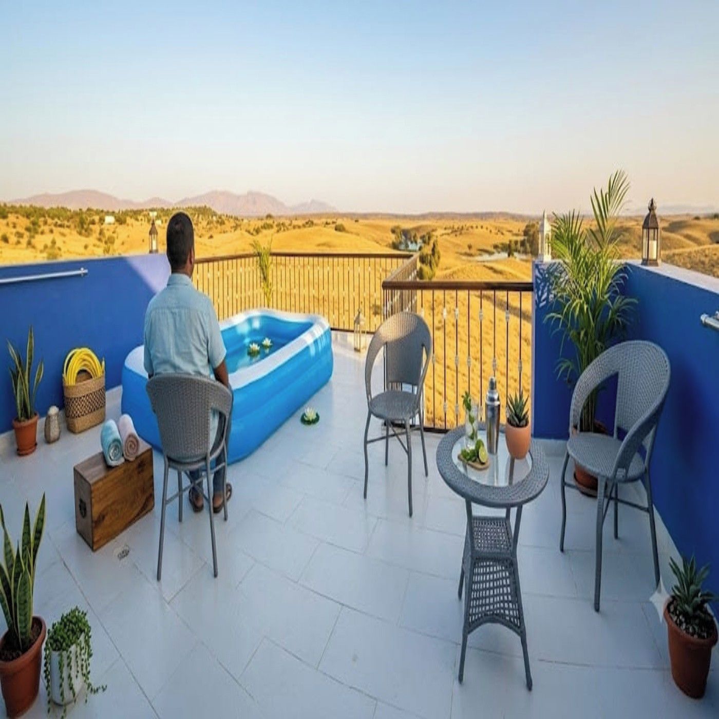 A man sitting on a balcony with a pool, surrounded by chairs and potted plants, overlooking a desert landscape.