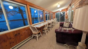 A room with a long table and chairs, large windows, a leather sofa, and a lamp. - Camp Come Again on Silver Lake–Pet-Friendly w/ Private Dock, Swimming & Views (Silver Lake)