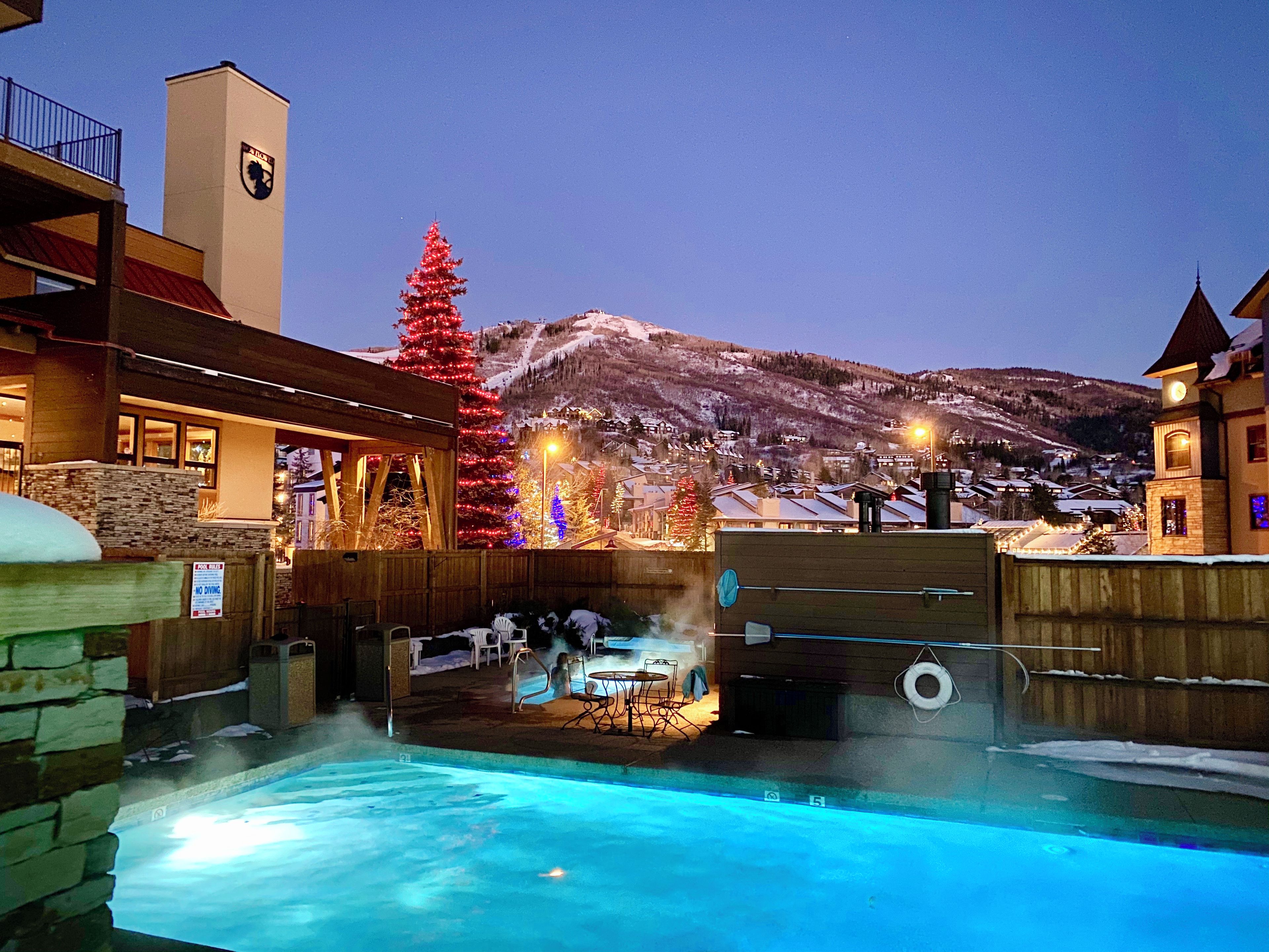 Un espace piscine extérieur chauffé, doté d’un jacuzzi, entouré de clôtures en bois et de murs en pierre, le tout se dressant sur un fond de montagnes enneigées et sous un ciel clair du soir.