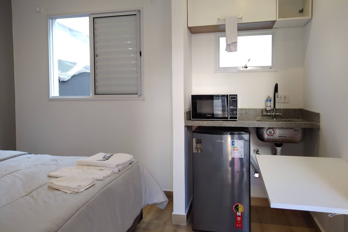Une chambre compacte comprenant un lit, une kitchenette équipée d’un four à micro-ondes et d’un réfrigérateur, ainsi qu’une fenêtre avec des stores.