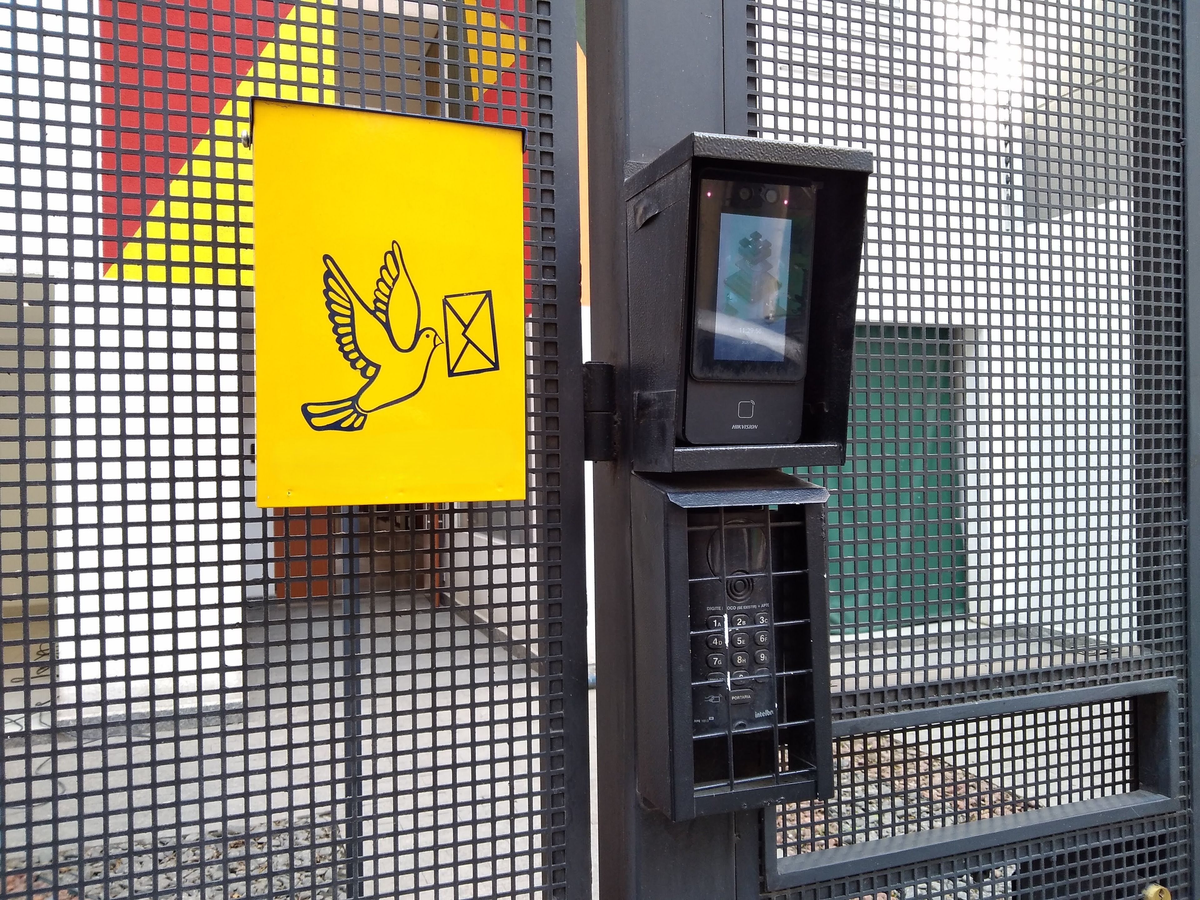 Une caméra de surveillance est fixée à une clôture métallique, et un panneau jaune représentant une colombe et une enveloppe est placé à proximité.