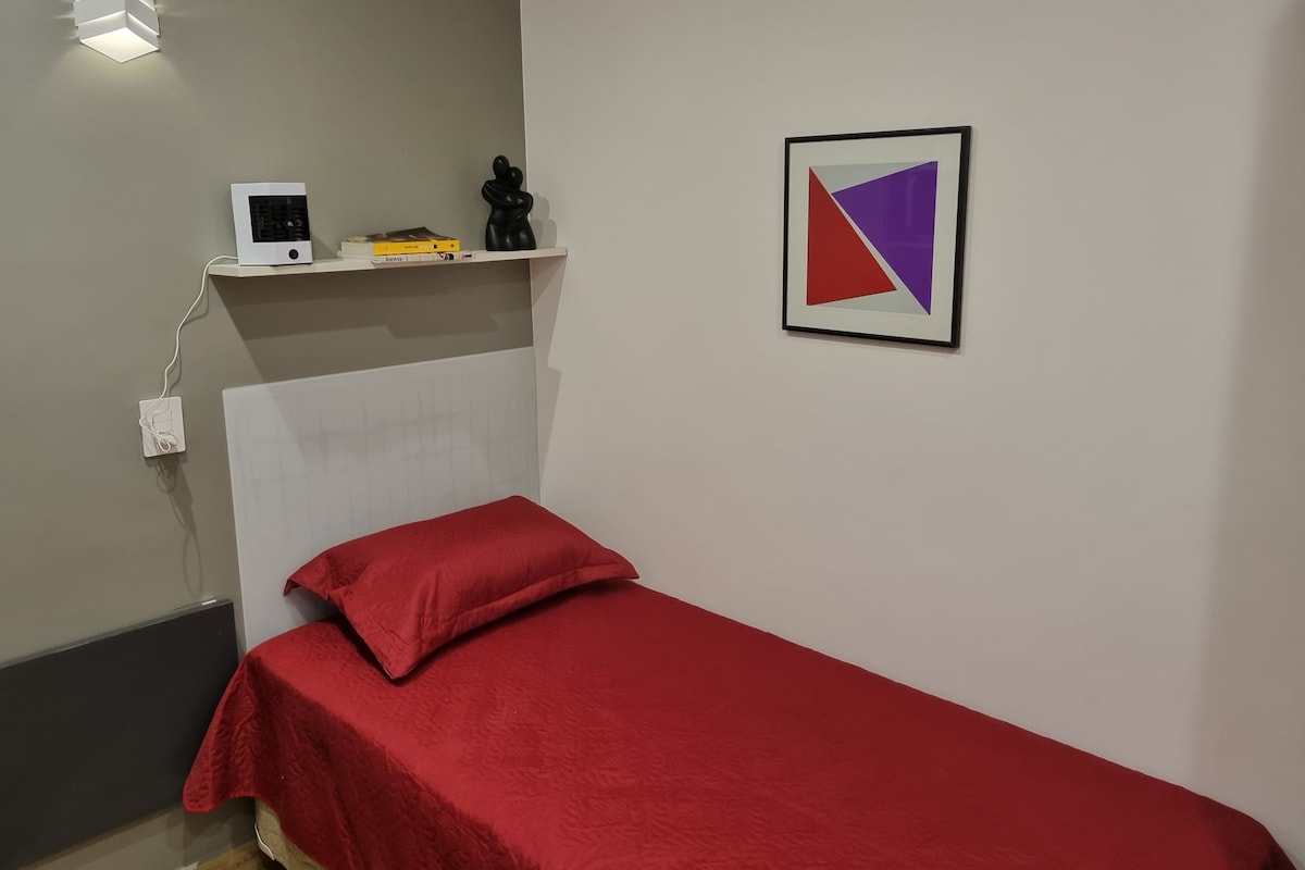 A small, single-bed room with a red bedspread, a white shelf with a small heater and books, and a framed geometric artwork on the wall.