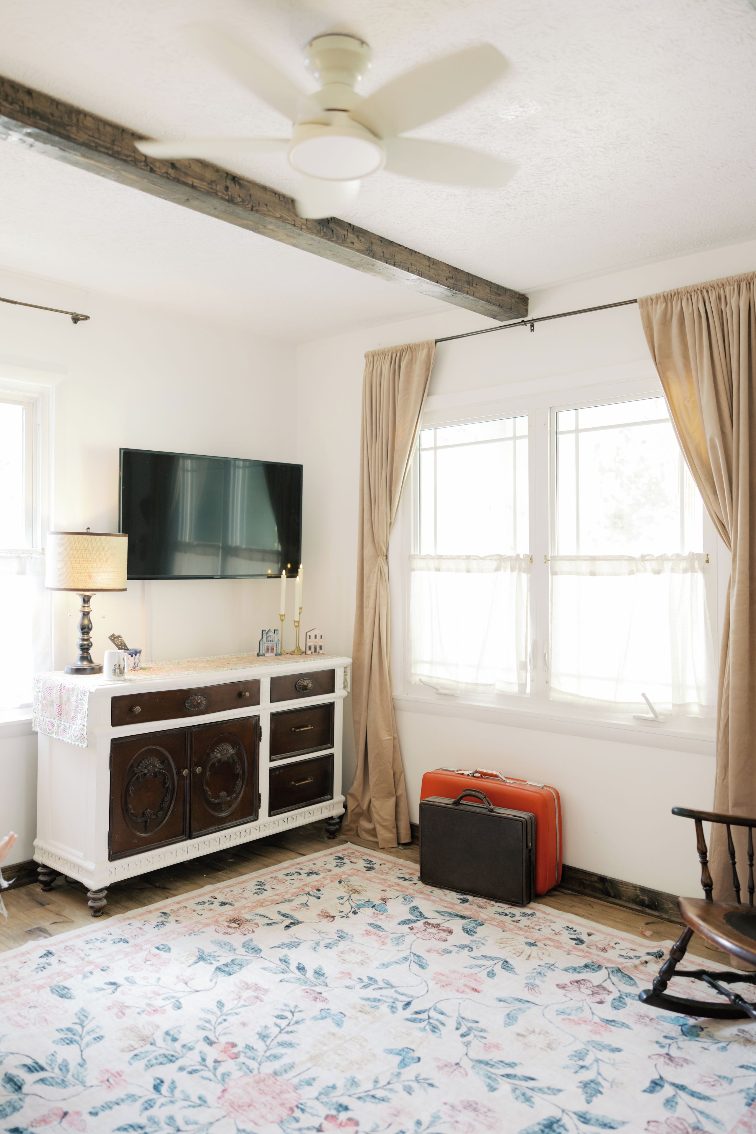 Een kamer met een plafondventilator, een flatscreen televisie, een dressoir in vintage stijl, een bloemenkleed en een koffer.