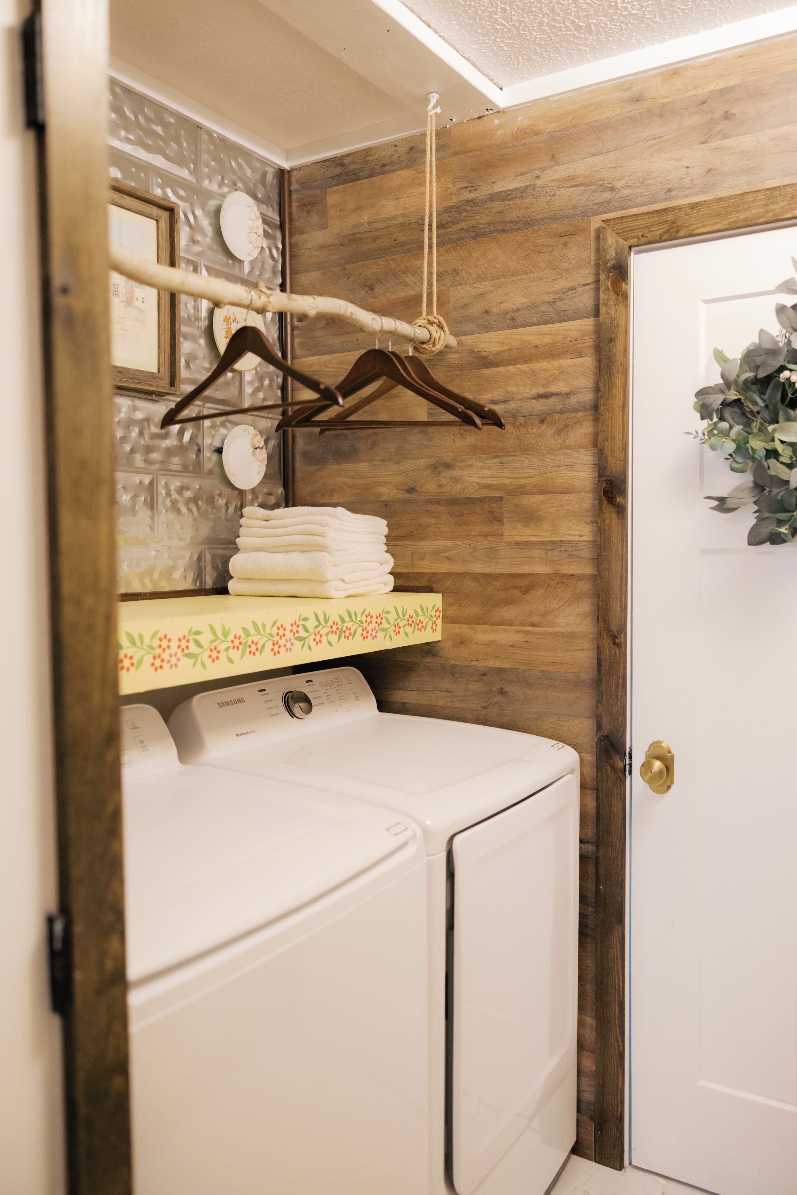 A laundry area with a white washing machine, a wooden shelf with folded towels, and a door with a plant decoration.