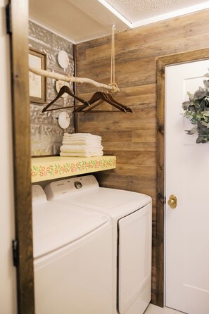 Un'area lavanderia con una lavatrice bianca, uno scaffale di legno con asciugamani piegati e una porta decorata con un motivo floreale.