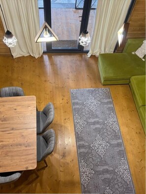 A room with a wooden floor, a green sofa, a patterned rug, and a dining table with chairs.