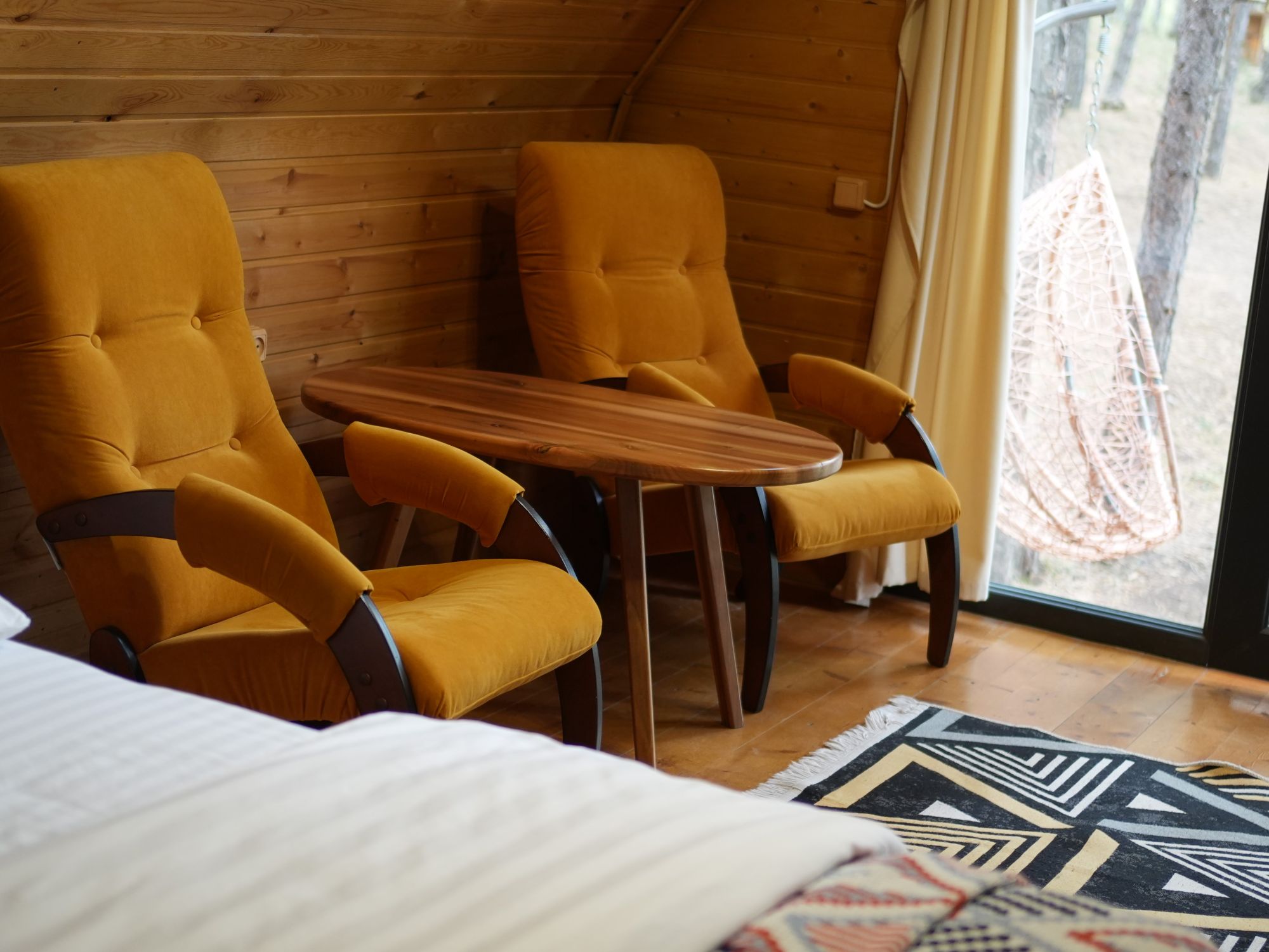 A cozy room with wooden walls, a small wooden table, two yellow armchairs, and a view of the outdoors through a window.