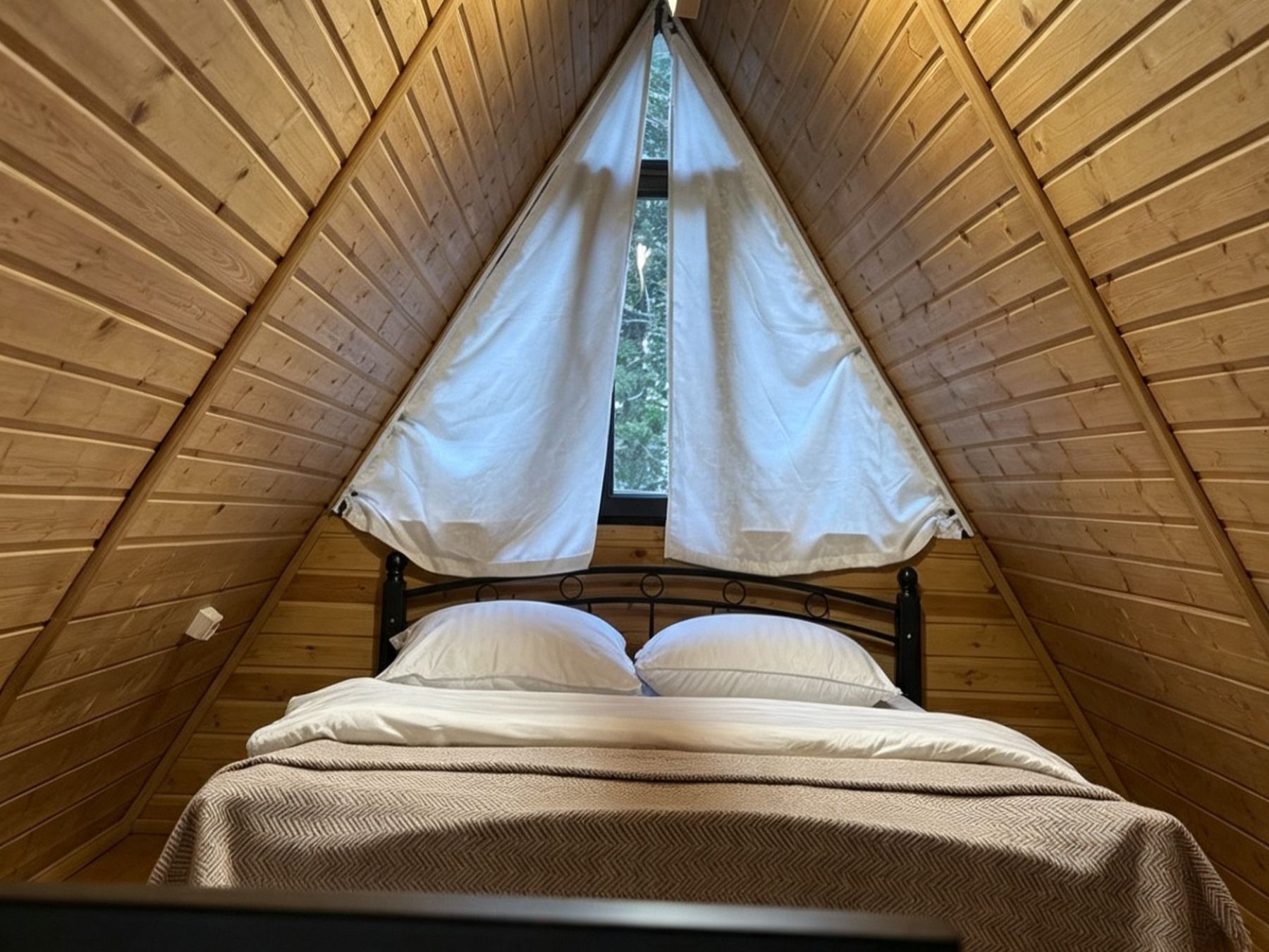 Eine dreieckige Holzzielung mit einem Bett in der Mitte, das einen Metallrahmen und weiße Bettwäsche hat.